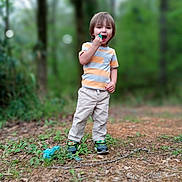 Damian is registered to the contest to win money with this photo: blue_object, boy, casual_clothing, child, daylight, forest, greenery, happy, nature, outdoor, pants, pathway, person, playing, smiling, sneakers, standing, striped_shirt, trees, young_child