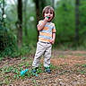 blue_object, boy, casual_clothing, child, daylight, forest, greenery, happy, nature, outdoor, pants, pathway, person, playing, smiling, sneakers, standing, striped_shirt, trees, young_child