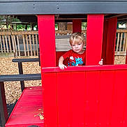 Tucker is registered to the contest to win money with this photo: child, playground, red, playhouse, wood_chips, fence, outdoor, blonde_hair, toy, window, wood, seat, casual_clothing, daylight, nature, tree, curious, young_child, play, park