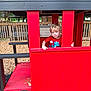 child, playground, red, playhouse, wood_chips, fence, outdoor, blonde_hair, toy, window, wood, seat, casual_clothing, daylight, nature, tree, curious, young_child, play, park