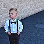 child, boy, toddler, suspenders, bow_tie, white_shirt, black_pants, black_shoes, standing, outdoor, brick_wall, sidewalk, fashion, portrait, casual, young_child, looking_away, serious, cute, small