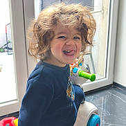 Kassim a rejoint le concours — aidez-le/la à gagner de superbes lots ! child, toddler, curly_hair, playful, tongue_out, indoor, window, toy, ride_on_toy, smiling, blue_clothing, floor, marble_floor, white_toy, green_handle, red_part, bright_light, person, happy, young_child