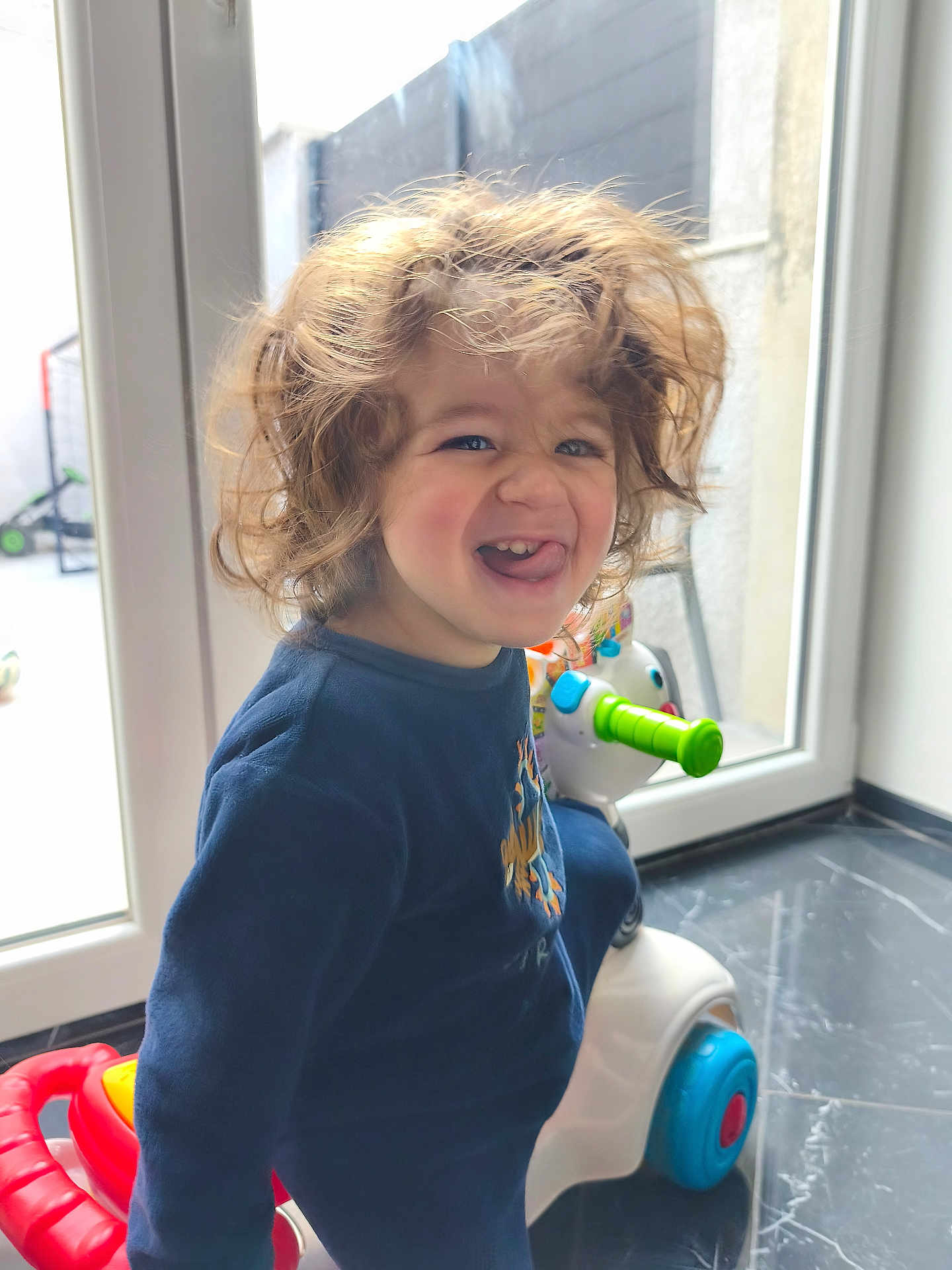 Kassim a rejoint le concours — aidez-le/la à gagner de superbes lots ! child, toddler, curly_hair, playful, tongue_out, indoor, window, toy, ride_on_toy, smiling, blue_clothing, floor, marble_floor, white_toy, green_handle, red_part, bright_light, person, happy, young_child