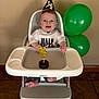 baby, high_chair, party_hat, cupcake, candle, balloon, celebration, smiling, infant, clothing, foot, face, hand, table, indoor, person, milestone, happy, head, birthday