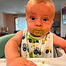 baby, child, high_chair, bib, green_food, messy, face, hand, indoor, curious, meal, feeding, clothing, expression, person, toddler, cute, food, smear, table
