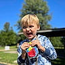 child, boy, happy, smiling, drink, juice_pouch, hoodie, blue_sky, outdoor, daylight, tree, greenery, casual_clothing, leisure, playful, portrait, person, fun, sunlight, nature