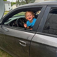 Eli is registered to the contest to win money with this photo: child, toddler, car, window, smiling, curly_hair, blue_shirt, outdoor, grass, steering_wheel, vehicle, door, suburban, person, happy, face, daylight, residential_area, automobile, joy
