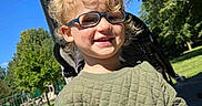 Eli joined the competition — help win amazing prizes! child, smiling, glasses, blonde_hair, curly_hair, green_sweater, outdoor, sunny, daylight, stroller, park, trees, grass, sidewalk, happy, person, portrait, young_child, casual_clothing, nature