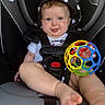 baby, child, car_seat, toy, colorful_ball, blue_eyes, hood, black_clothing, white_hearts, smiling, tongue_out, cute, infant, seatbelt, indoor, closeup, chubby_cheeks, playful, person, portrait