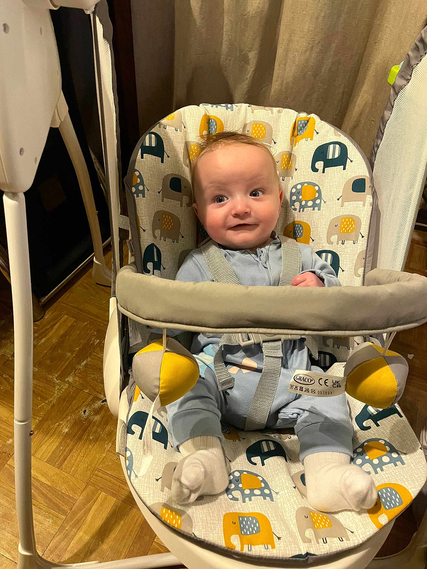 Caleb is registered to the contest to win money with this photo: baby, infant, baby_swing, elephant_pattern, toy, indoor, floor, clothing, white_socks, straps, seat, curtain, wood_floor, play, child, person, cute, smile, expression, sitting