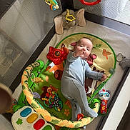 Caleb is registered to the contest to win money with this photo: baby, playpen, toy, onesie, infant, plush_toy, colorful, floor_mat, rattle, piano_toy, animal_design, curious, child, indoor, soft_toys, hand_reaching, lying_down, playtime, cute, young_child