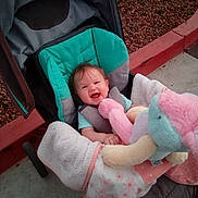 Esme is registered to the contest to win money with this photo: baby, stroller, plush_toy, blanket, happy, smiling, child, outdoor, concrete, curb, gravel, person, clothing, head, seat, comfort, cute, infant, toy, joy