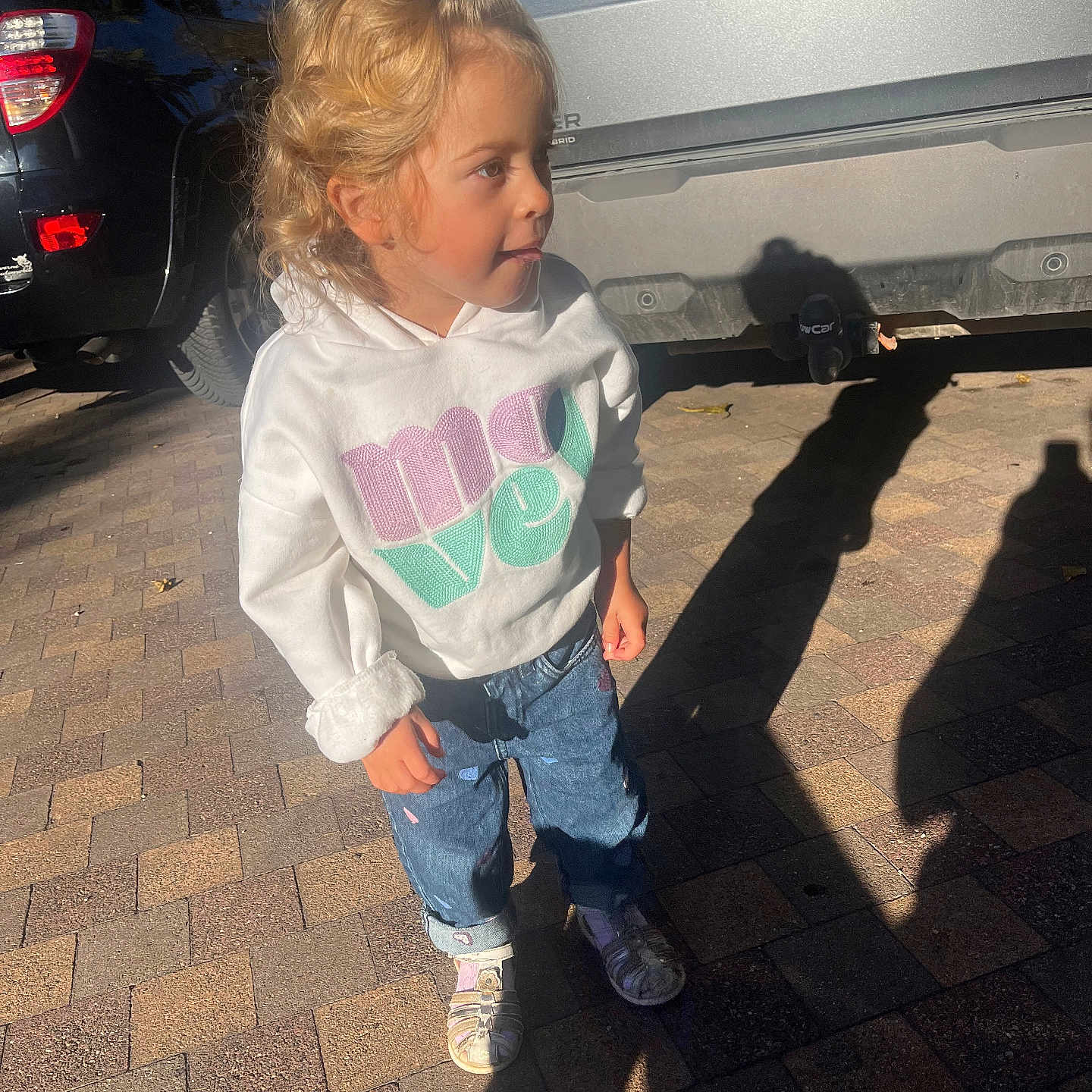 Iris participe au concours pour gagner de l'argent avec cette photo : blonde_hair, car, casual_clothing, child, curious_expression, curly_hair, daylight, hair_clip, hoodie, jeans, outdoor, pavement, person, shadow, shoes, side_view, small_child, standing, street, sunlight