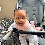 Isla is registered to the contest to win money with this photo: baby, child, stroller, pink_bib, hair_buns, curious_expression, indoor, water_bottles, gray_stroller, striped_clothing, playful, infant, seat, baby_clothes, toy_free, happy, cuteness, person, baby_face, baby_gear