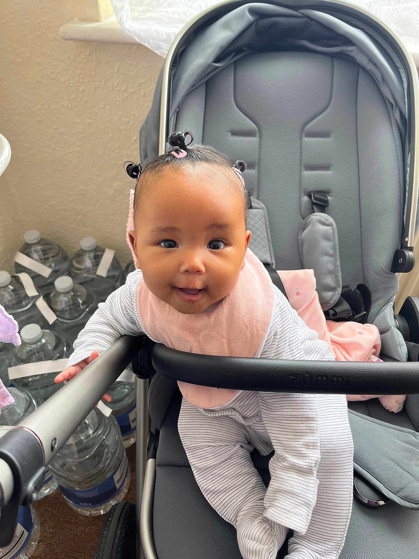 Isla is registered to the contest to win money with this photo: baby, child, stroller, pink_bib, hair_buns, curious_expression, indoor, water_bottles, gray_stroller, striped_clothing, playful, infant, seat, baby_clothes, toy_free, happy, cuteness, person, baby_face, baby_gear