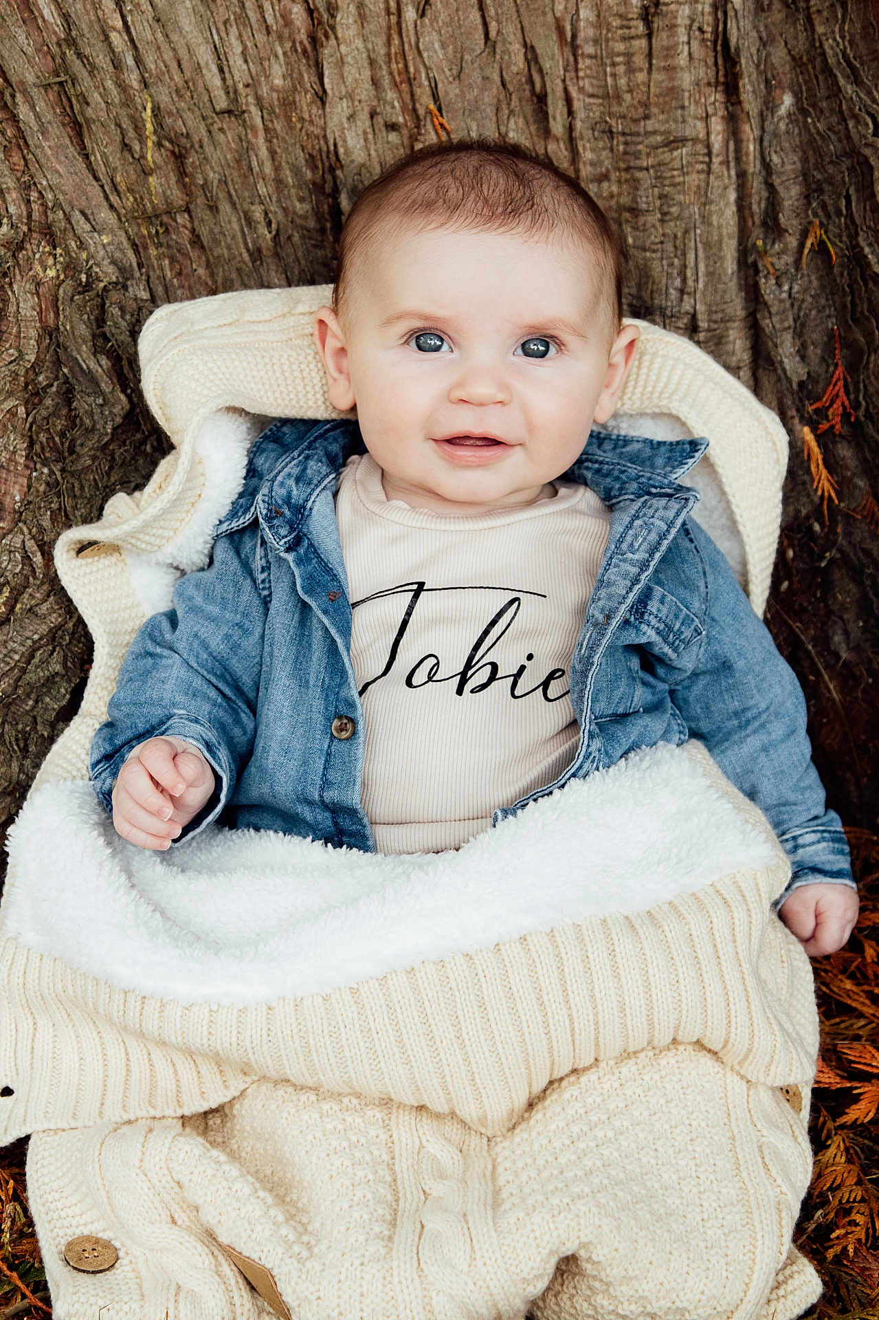 Tobie is registered to the contest to win money with this photo: baby, child, denim_jacket, knitwear, blanket, tree_trunk, outdoor, fall_leaves, smiling, face, portrait, cute, warm_clothing, sitting, infant, nature, closeup, happy, person, cozy