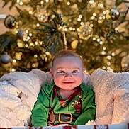 Tobie is registered to the contest to win money with this photo: baby, child, smile, christmas_tree, holiday, festive_clothing, blanket, cozy, lights, decorations, indoors, portrait, happy, cute, christmas, red, green, winter, toy, celebration