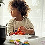 child, curly_hair, scissors, crafting, table, paper, indoor, light, curtains, sitting, concentration, clothing, hands, face, expression, toy_scissors, colorful, kid, activity, home