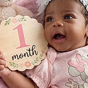 June is registered to the contest to win money with this photo: baby, infant, child, pink, headband, teddy_bear, plush_toy, blanket, sign, month, celebration, smiling, cute, clothing, portrait, hand, face, lying_down, soft_texture, happy