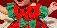 Sophia Lana is registered to the contest to win money with this photo: baby, child, watermelon_costume, red_hat, green_bow, smiling, sitting, bed, pillow, happy, cute, clothing, costume, indoors, infant, headwear, footwear, red_shoes, person, portrait