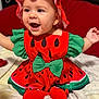 baby, child, watermelon_costume, red_hat, green_bow, smiling, sitting, bed, pillow, happy, cute, clothing, costume, indoors, infant, headwear, footwear, red_shoes, person, portrait