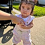 car, casual_clothing, child, curious_expression, daylight, grass, hand_holding, holding_hands, outdoor, overalls, person, pigtails, portrait, sidewalk, stroller, sunlight, toddler, urban, white_shoes, young_child