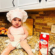 Teresa is registered to the contest to win money with this photo: child, toddler, chef_hat, apron, flour, kitchen, countertop, baking, messy, food, bread, whipping_cream, carton, cooking, face, hands, legs, cabinet, cutting_board, tile_backplash