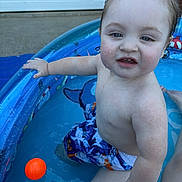 Caiden is registered to the contest to win money with this photo: baby, child, toddler, swimming, pool, water, outdoor, summer, wet_hair, swim_trunks, orange_ball, toy, sitting, portrait, face, smiling, curious, backyard, water_play, photography
