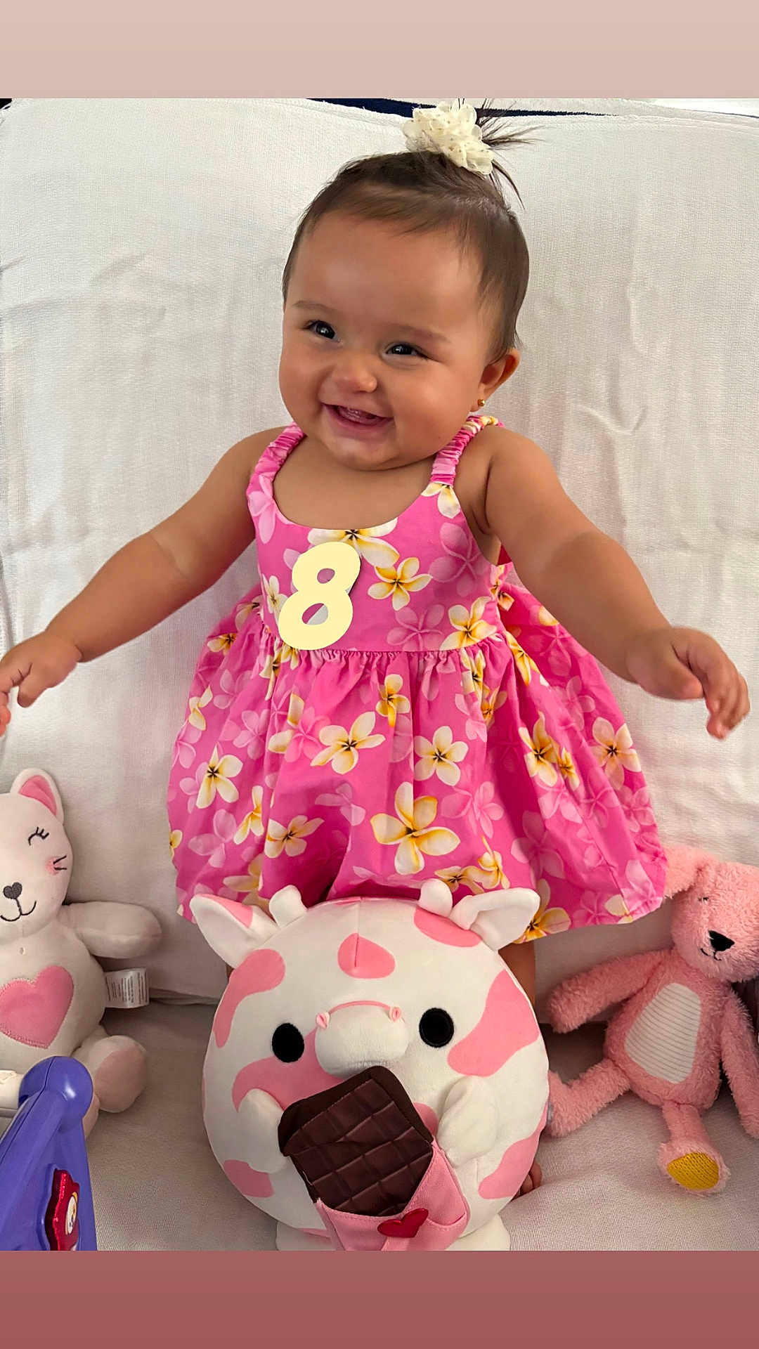 Milani joined the competition — help win amazing prizes! baby, child, smiling, pink_dress, floral_pattern, plush_toy, stuffed_animal, cat_toy, cow_toy, bunny_toy, white_blanket, happy, indoors, cute, sitting, person, face, toy, playful, colorful