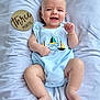baby, infant, smiling, romper, blue_clothing, bracelet, white_blanket, wooden_sign, three_months, cute, happy, lying_down, chubby_legs, portrait, child, indoors, soft_texture, closeup, face, skin