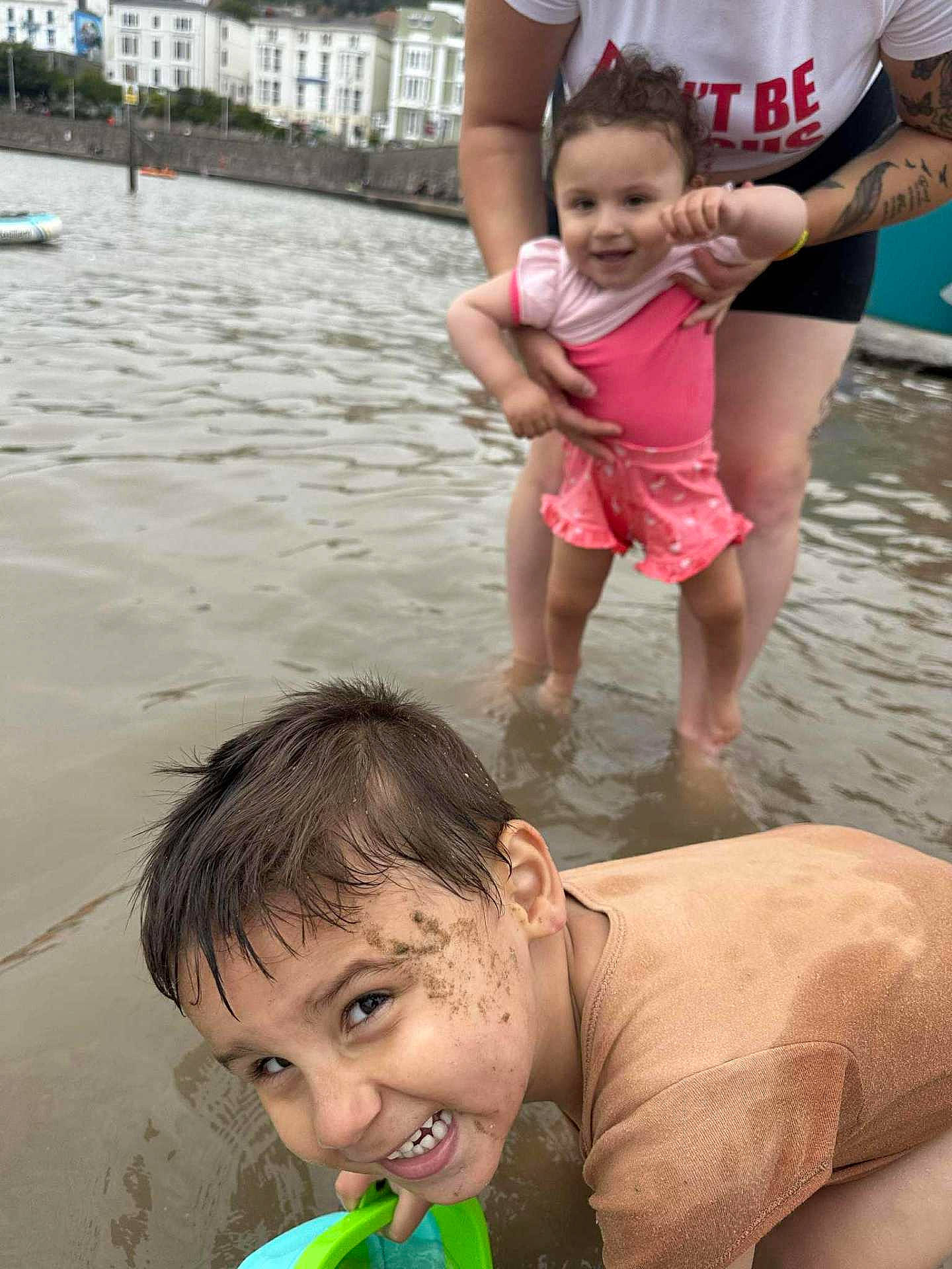 Armani-Rose is registered to the contest to win money with this photo: adult, bucket, buildings, child, daytime, family, fun, happy, mud, outdoor, person, play, shallow_water, shirt, shorts, smile, summer, tattoo, toddler, water
