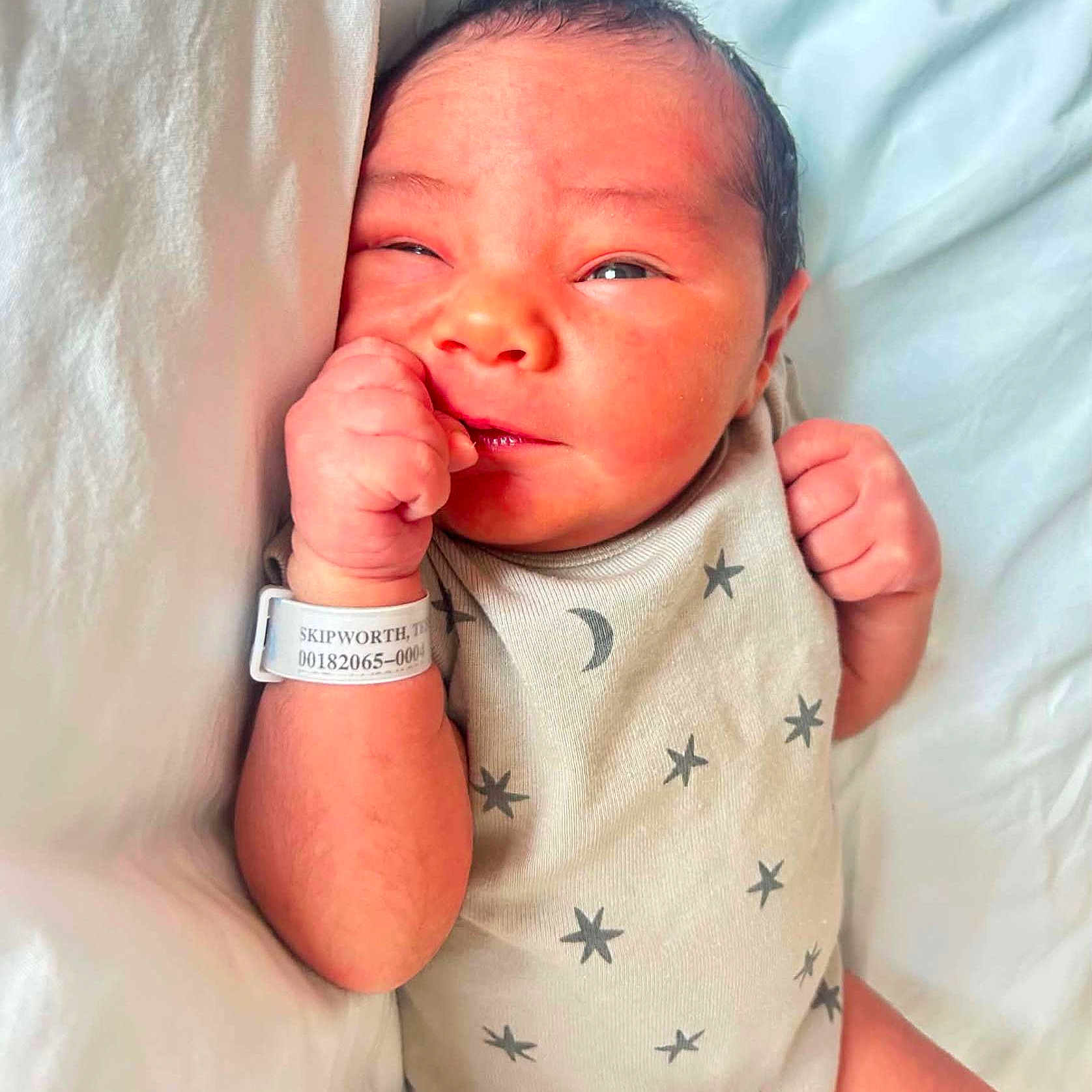 Zayden is registered to the contest to win money with this photo: newborn, baby, infant, onesie, stars, moon, hospital_bracelet, bed, blanket, sleepwear, cute, skin, hand, face, expression, portrait, resting, indoors, person, young_child