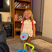 Novaleigh is registered to the contest to win money with this photo: backpack, blonde_hair, books, bookshelf, carpet, child, clothes, dog_bed, floor, happy, indoor, living_room, portrait, purse, rug, smiling, speaker, toy_cart, tv, wooden_shelf