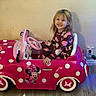 child, christmas_tree, electric_outlet, floor, girl, happy, holiday_lights, indoor, minnie_mouse, pajamas, pink, plug, polka_dot, seat, smile, steering_wheel, toddler, toy, toy_car, wall