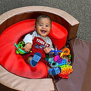 Jace is registered to the contest to win money with this photo: baby, carpet, colorful_toys, cute, foam_cushion, happy, indoor_playroom, infant, onesie, play_seat, pop_it, portrait, rattle, red_shirt, sitting, smiling, socks, teething_toy, toddler, toy