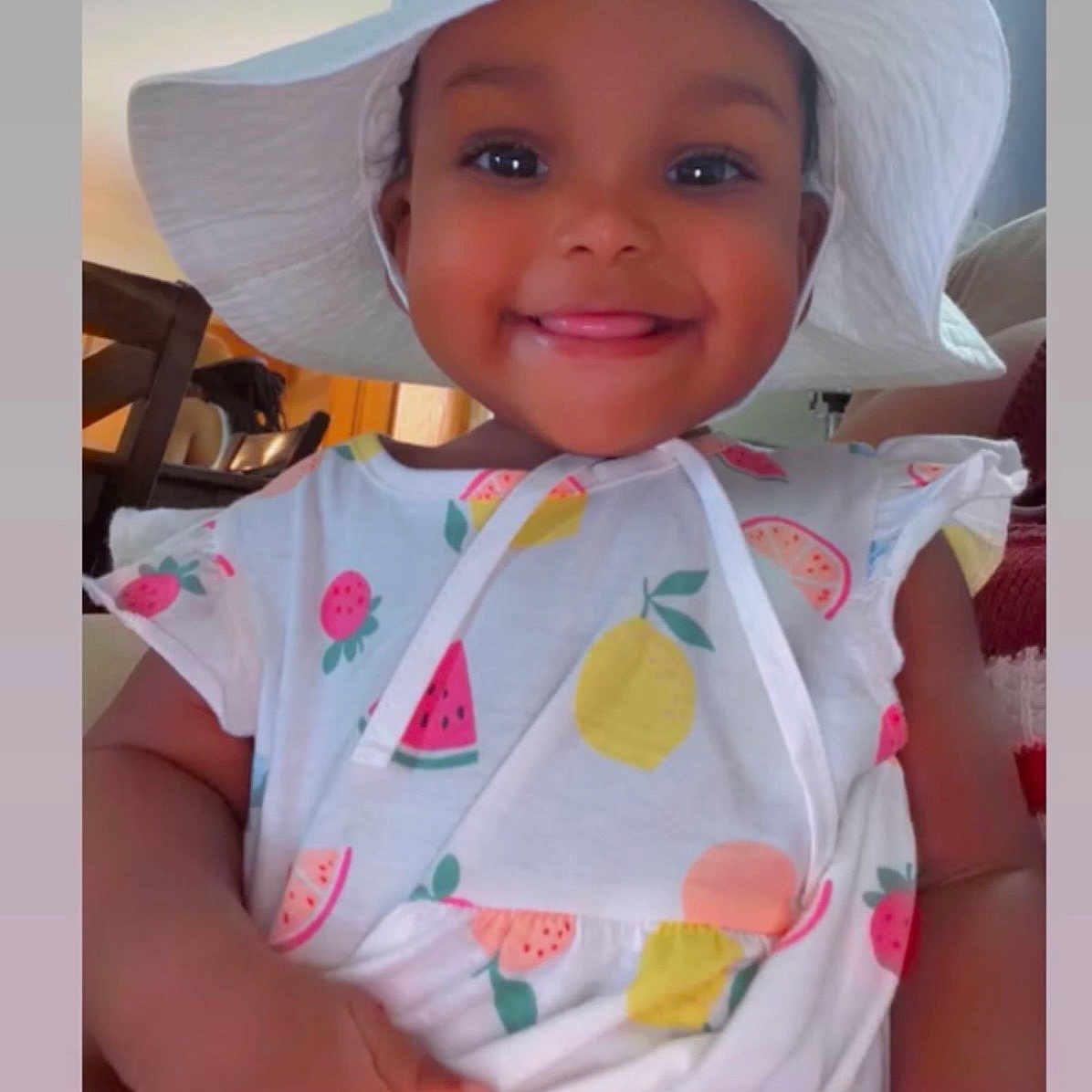 Alora joined the competition — help win amazing prizes! toddler, child, smiling, hat, white_hat, fruit_pattern, dress, indoor, furniture, happy, cute, portrait, person, young_child, skin_tone, hand, tongue_out, colorful_clothing, seated, cozy