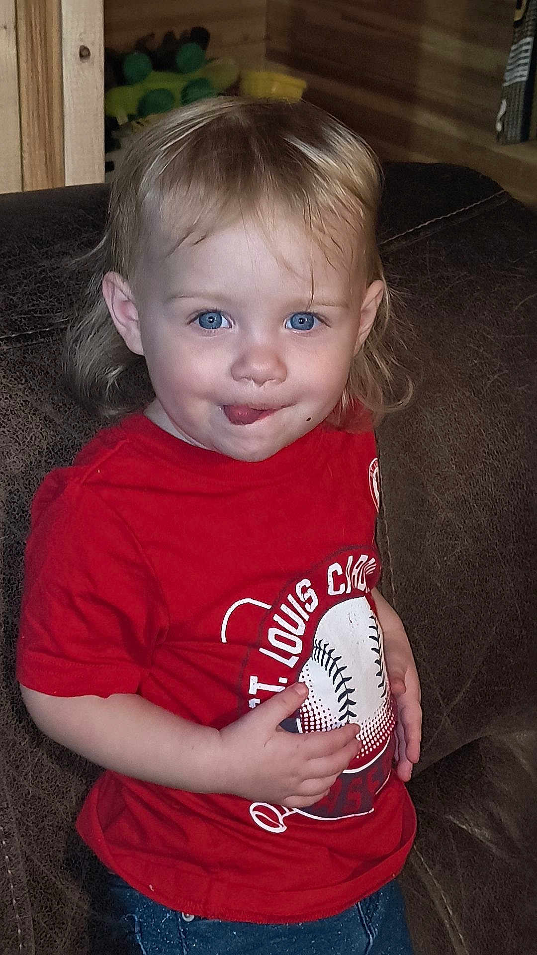 Kendalynn is registered to the contest to win money with this photo: toddler, child, baby, blue_eyes, red_tshirt, baseball, st_louis_cardinals, jeans, couch, leather, indoor, portrait, smile, mischievous, cute, face, hair, hand, person, sitting