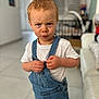 toddler, child, denim_overalls, white_shirt, indoor, portrait, serious_expression, blond_hair, short_hair, standing, floor_tiles, furniture, sofa, baby, person, cute, closeup, home, young_child, casual_clothing