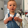 toddler, child, denim_overalls, white_tshirt, indoor, curly_hair, expressive_face, hands, person, young_child, home_interior, couch, blurred_background, portrait, cute, casual_clothing, curious_expression, light_skin, standing, necklace