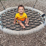 Kylo is registered to the contest to win money with this photo: toddler, child, swing, rope_swing, playground, orange_shirt, smiling, outdoor, wood_chips, greenery, happy, baby, person, seat, net, summer, fun, cute, park, leisure