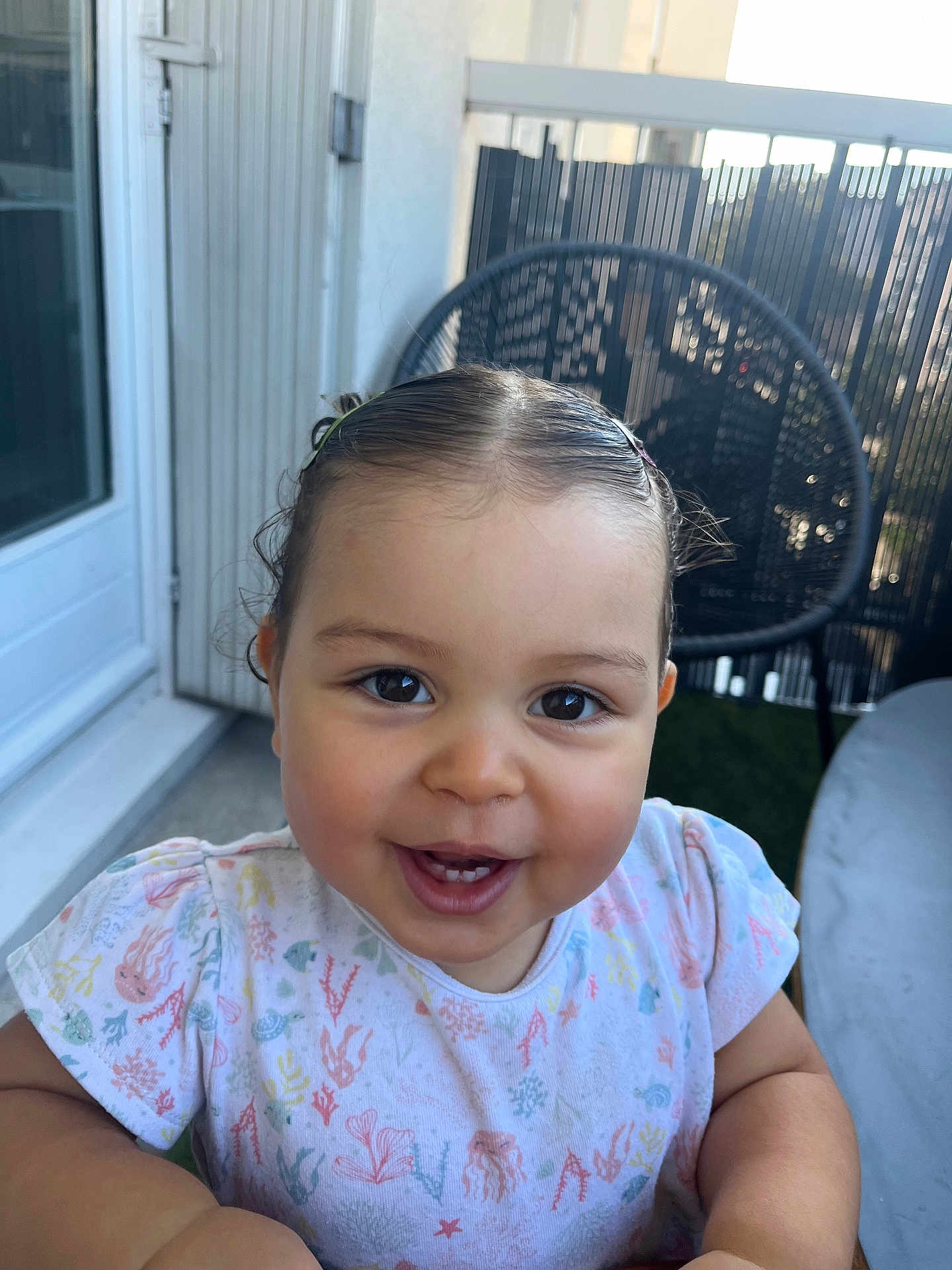 Mina participe au concours pour gagner de l'argent avec cette photo : toddler, child, smile, happy, face, hair, shirt, patterned_clothing, outdoor, chair, table, fence, balcony, daylight, person, cute, baby, skin, expression, sitting