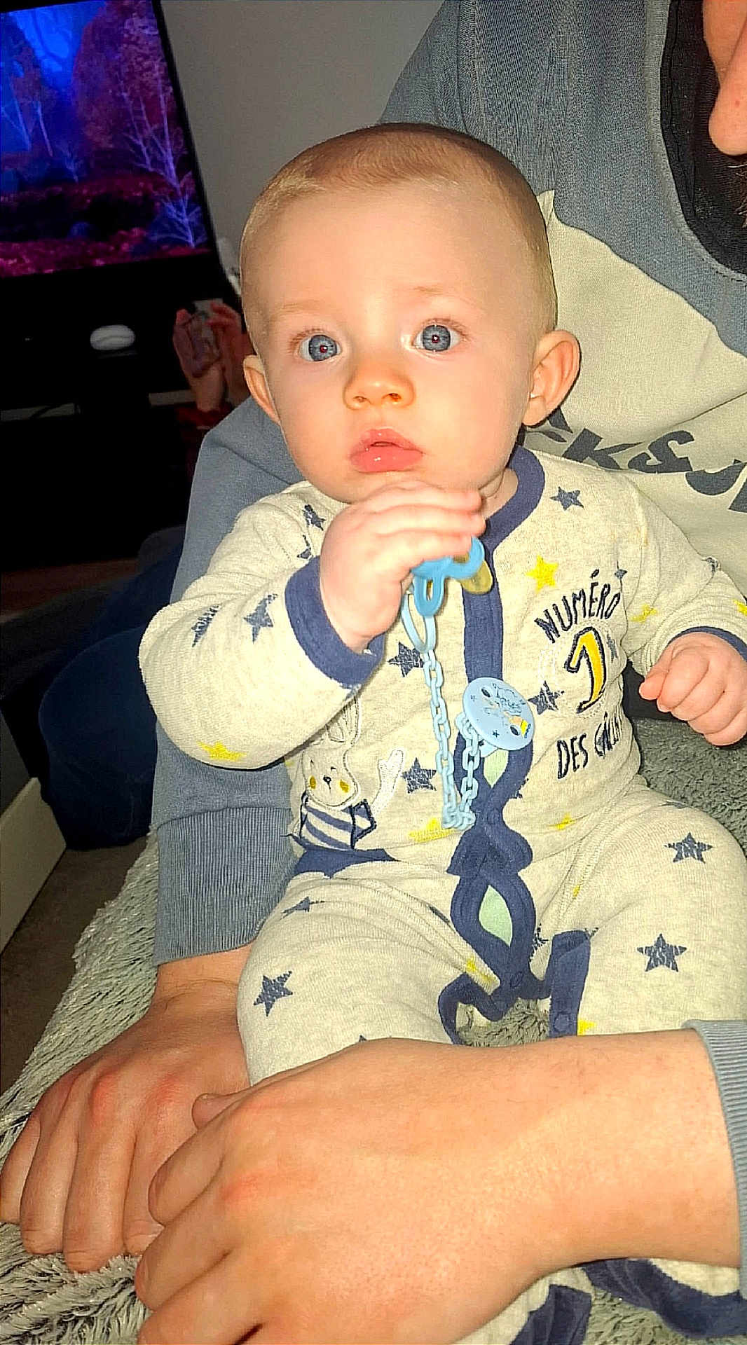 Ayden a rejoint le concours — aidez-le/la à gagner de superbes lots ! baby, child, pacifier, pajamas, indoor, person, hand, adult, living_room, television, furniture, blue_eyes, comfort, cute, holding, sitting, star_pattern, clothing, closeup, portrait