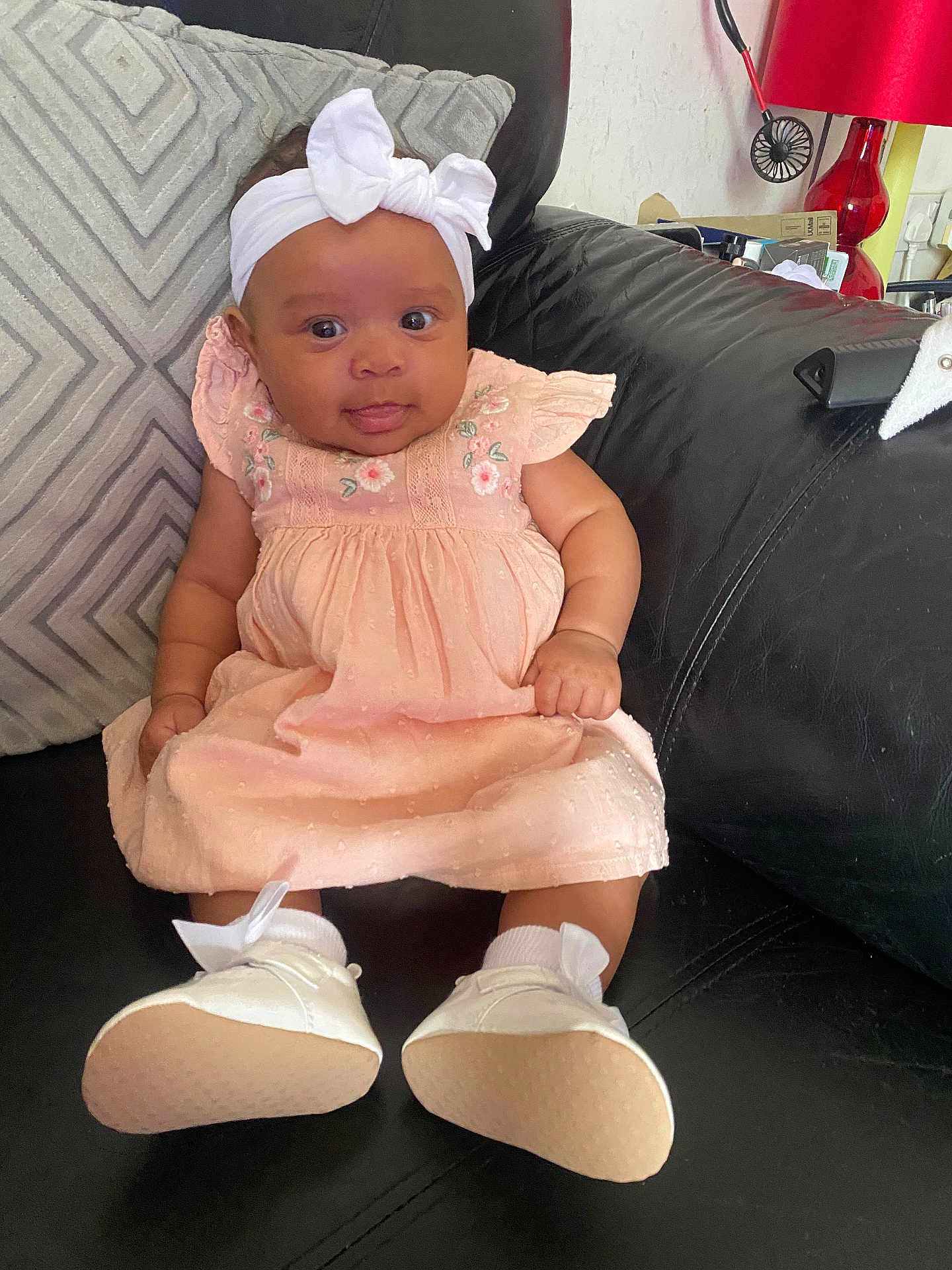 Amara-Mae is registered to the contest to win money with this photo: baby, child, infant, girl, dress, headband, white_bow, shoes, socks, couch, pillow, black_leather, home_interior, lamp, red_lamp, curious_expression, seated, indoor, cute, portrait