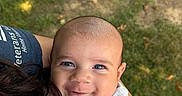 Matthias is registered to the contest to win money with this photo: baby, child, infant, smiling, face, blue_eyes, outdoor, grass, nature, portrait, close_up, cuddling, parent, adult, arms, gray_clothing, long_sleeves, sunlight, happy, family