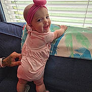 Stormi joined the competition — help win amazing prizes! baby, blanket, bow, child, couch, footwear, hand, happy, headband, home, indoor, infant, person, pink_dress, smiling, standing, support, tattoo, tropical_pattern, window_blinds