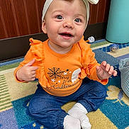 Everlee joined the competition — help win amazing prizes! baby, child, smiling, orange_shirt, headband, bow, sitting, carpet, indoor, happy, cute, white_socks, blue_pants, floor, toy, wooden_wall, infant, clothing, person, portrait