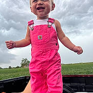 Brianna is registered to the contest to win money with this photo: toddler, child, pink_overalls, smiling, outdoor, cloudy_sky, truck_bed, green_field, happy, person, clothing, nature, grass, daylight, portrait, cute, young_child, standing, casual, fun