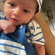 Thelio participe au concours pour gagner de l'argent avec cette photo : baby, infant, hat, striped_clothing, blue_overalls, buttons, hand, arm, face, eyes, portrait, closeup, smile, skin, fabric, bedding, person, newborn, cute, indoor