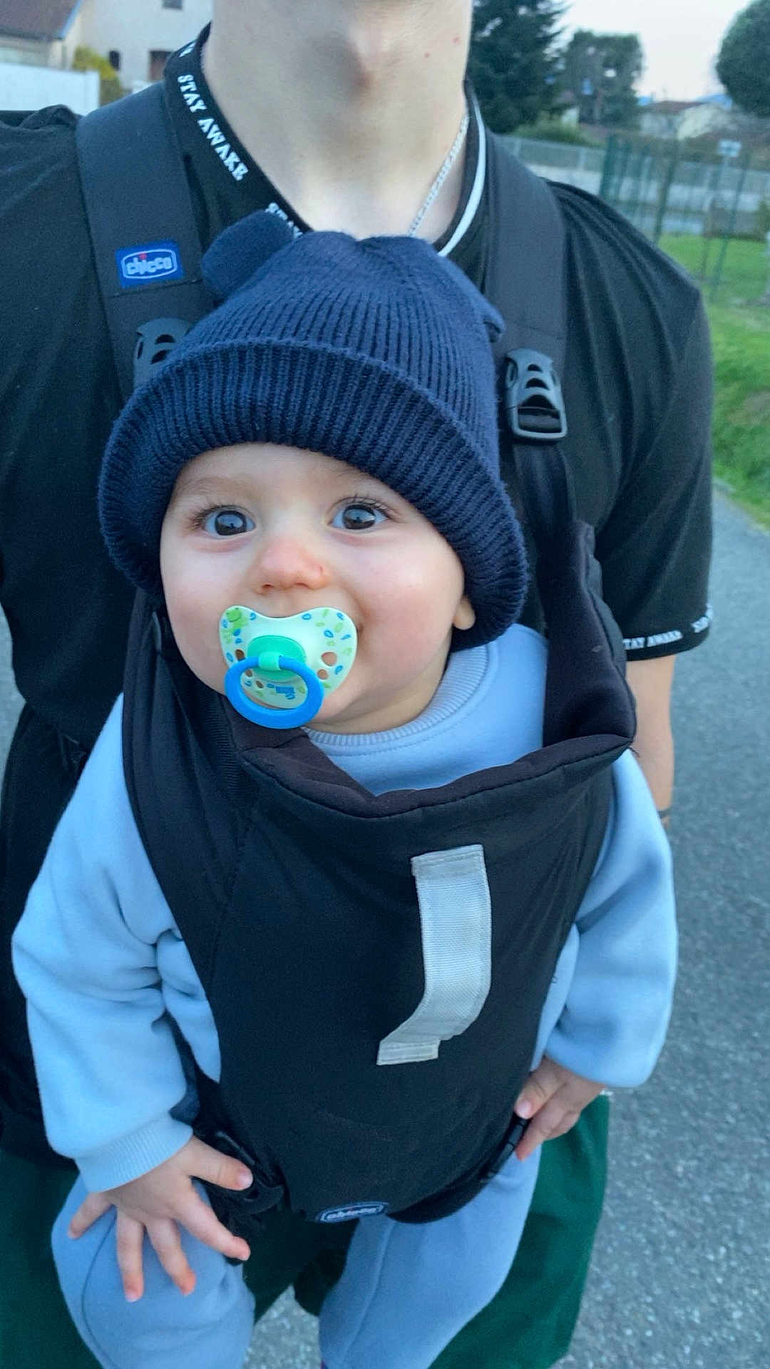 Thelio participe au concours pour gagner de l'argent avec cette photo : baby, pacifier, navy_beanie, knit_hat, baby_carrier, carrier, adult_torso, black_shirt, big_eyes, cute, smiling, hands, sweater, outdoors, sidewalk, grass, portrait, close_up, blue_pacifier, infant_clothing