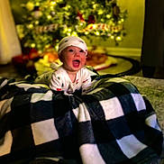 Matt joined the competition — help win amazing prizes! baby, infant, child, christmas_tree, holiday_lights, blanket, plaid, onesie, hat, smile, happy, living_room, bokeh, toy_train, carpet, curtains, sofa, portrait, indoors, decorations